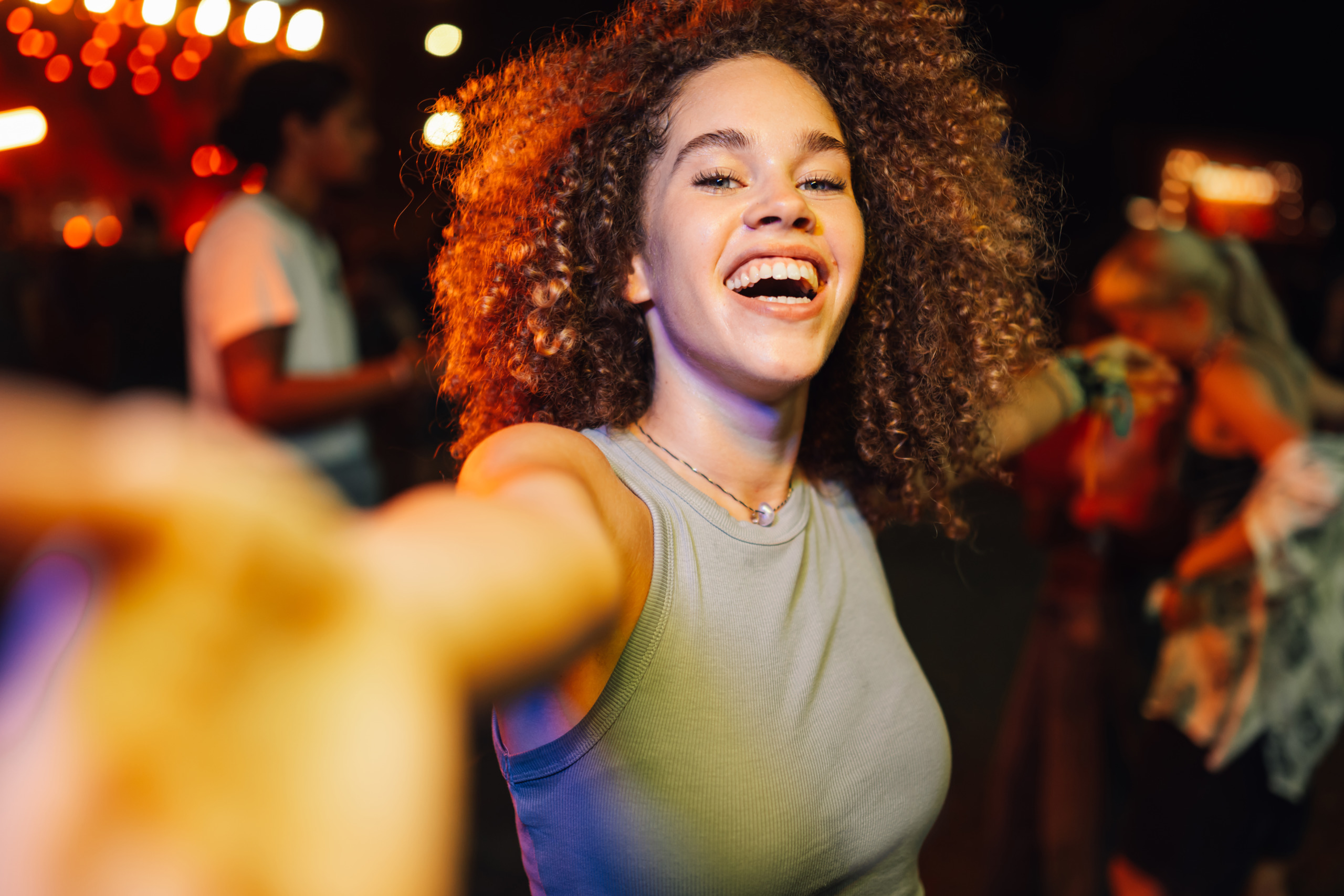Young woman enjoying live music and reaching for the camera at night concert Enthusiastic young woman with curly hair enjoying live music performance, reaching out to the camera while dancing with friends at night concert, creating a sense of shared excitement and joy